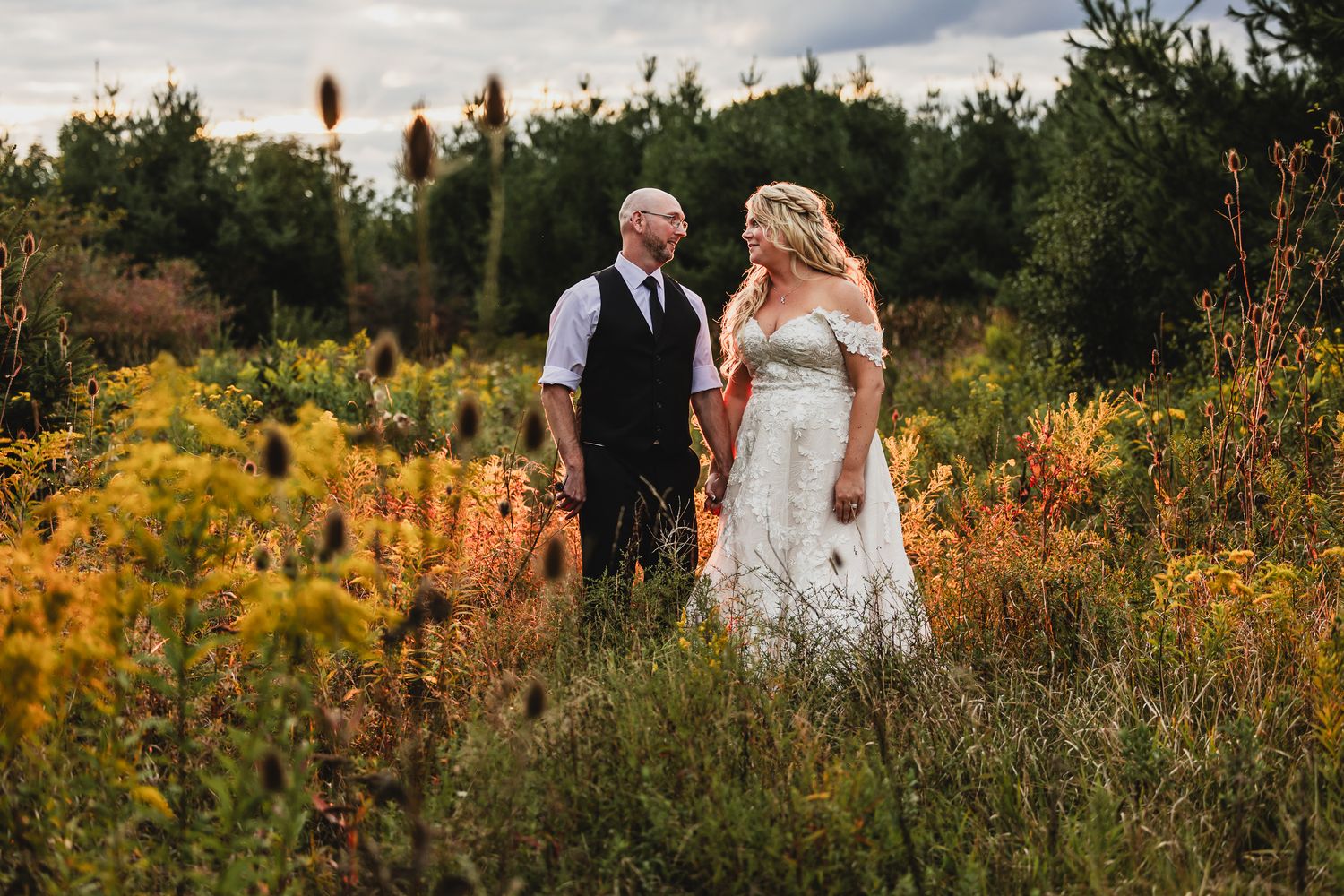 A couple walks through a vibrant autumn meadow with golden flowers and tall grass at sunset.