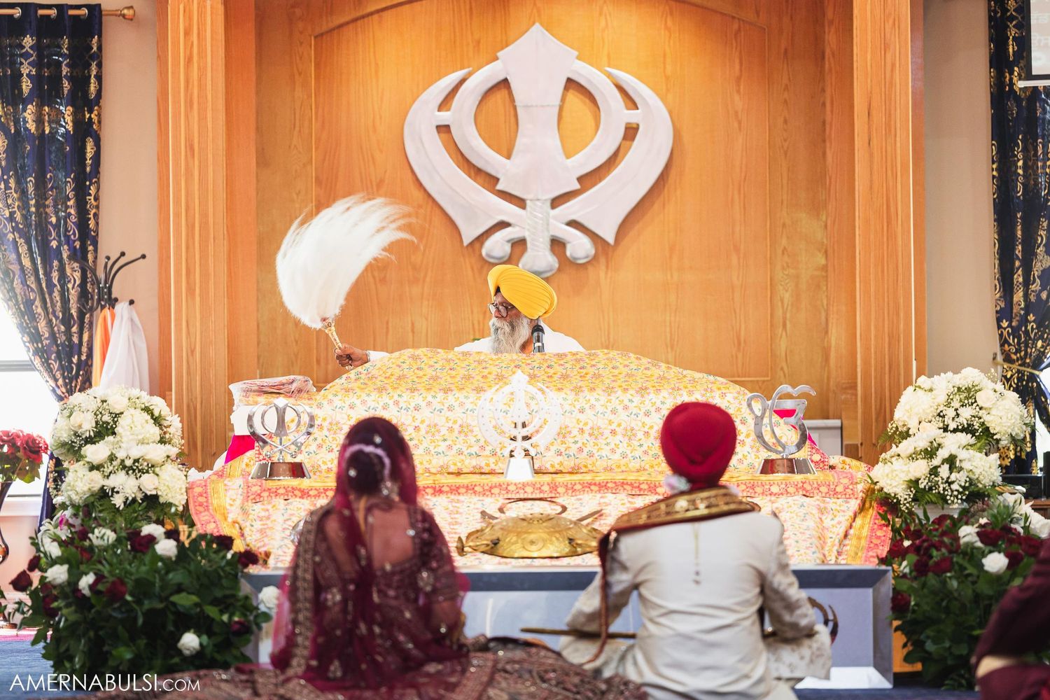 Sikh wedding ceremony in front of altar with Khanda symbol and floral decorations.