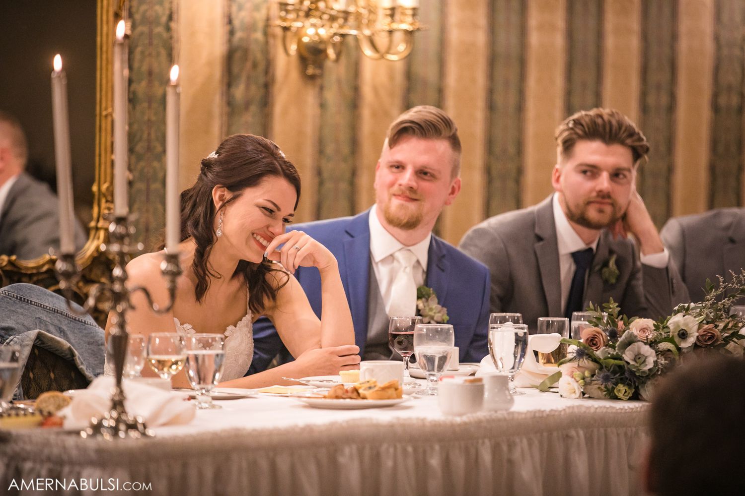 Wedding reception guests seated at elegant tables with chandeliers and floral centerpieces in a grand banquet hall.