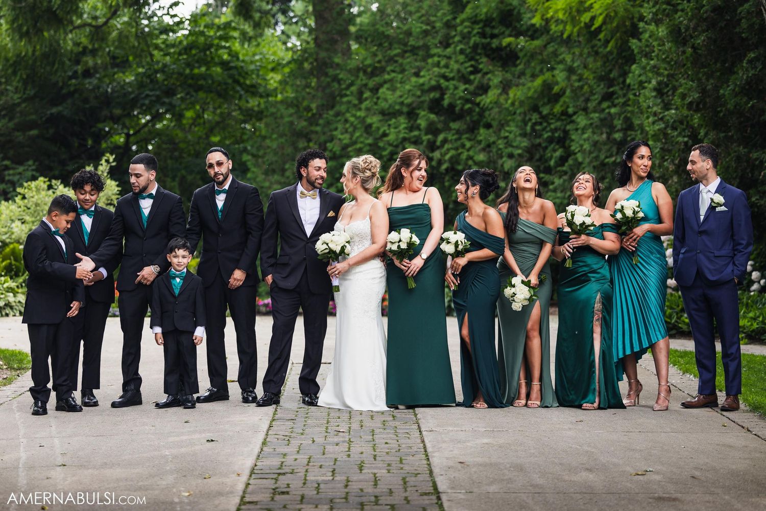 Wedding party lines up on garden path with emerald green bridesmaid dresses and black suits.