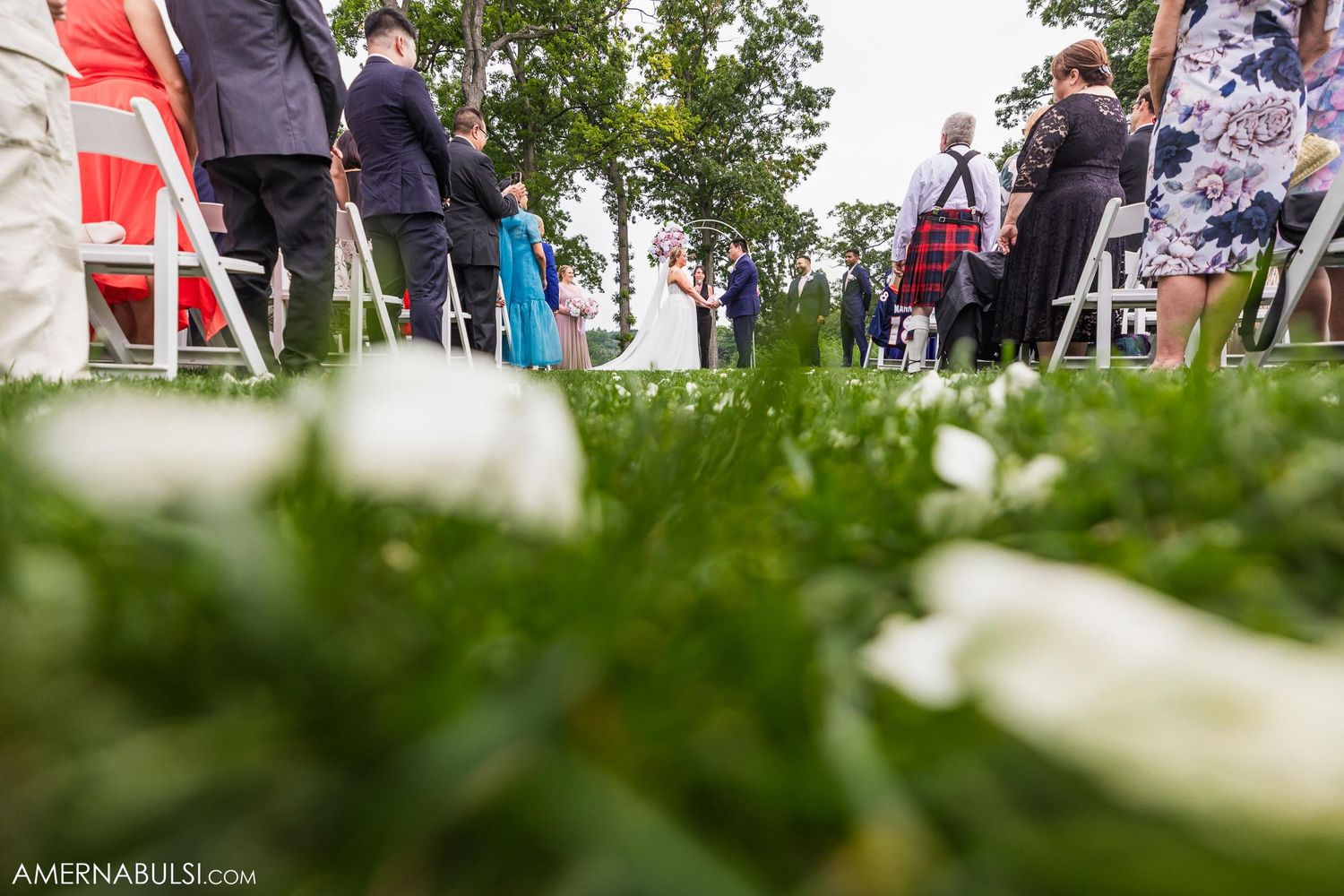 Low angle view of wedding ceremony through grass with guests' feet visible in background.