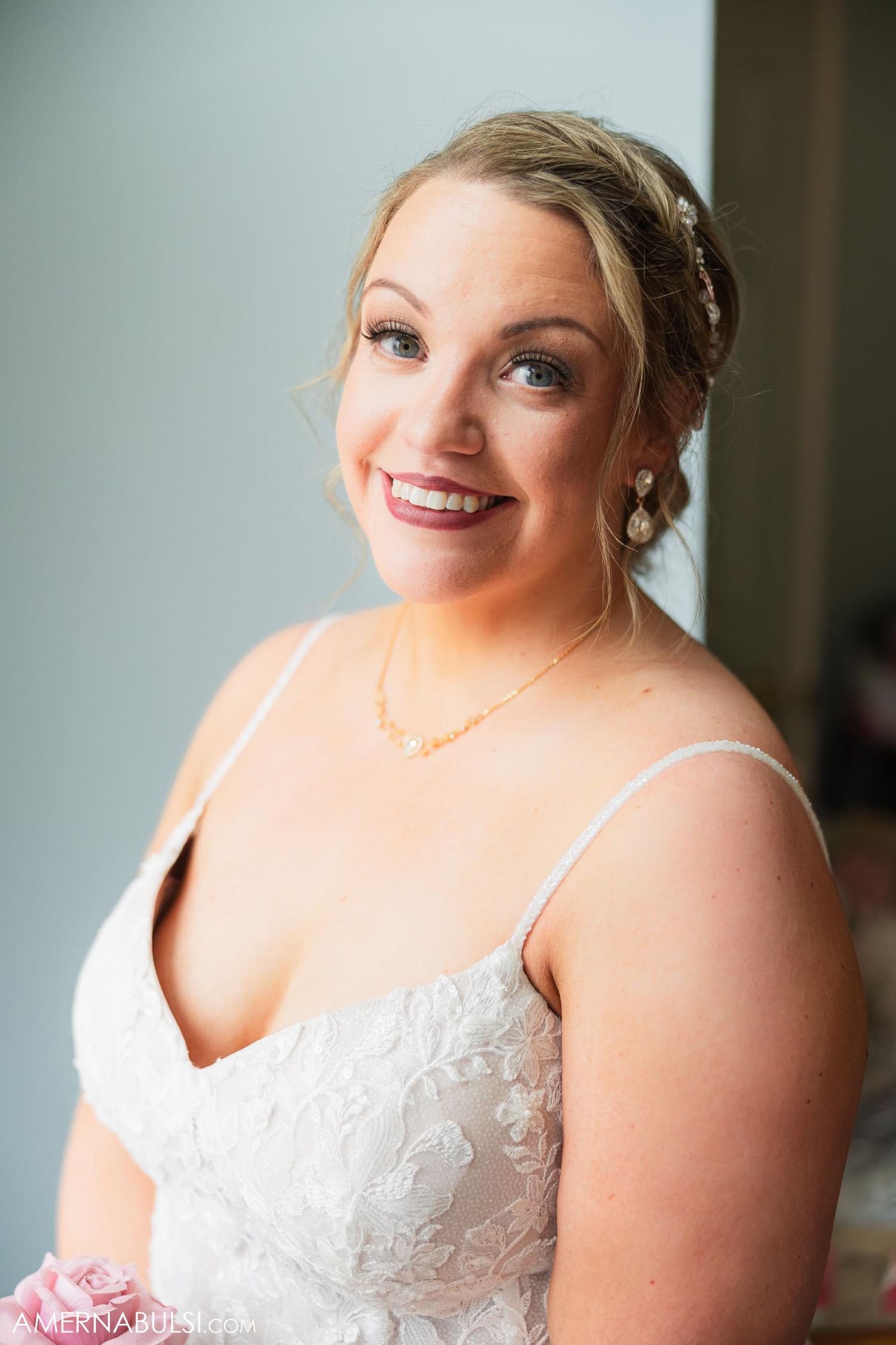 A bride in a white lace wedding dress smiles warmly while wearing delicate jewelry.