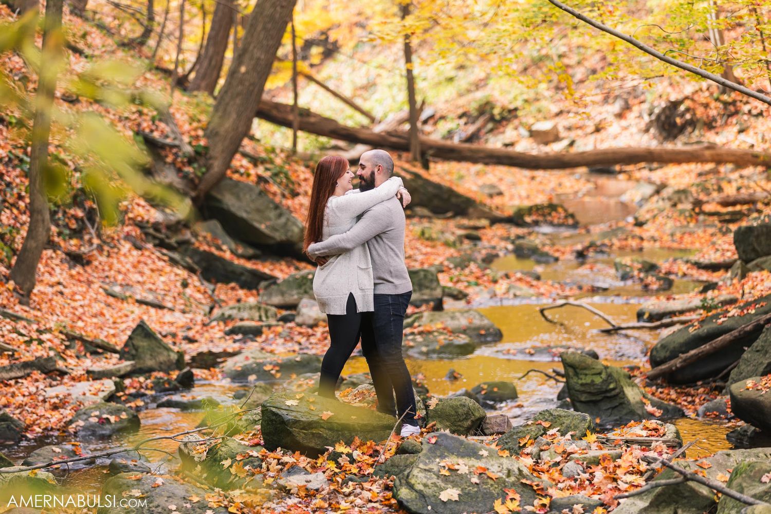 Couple hugging on rocky creek bed with orange fall foliage in background.