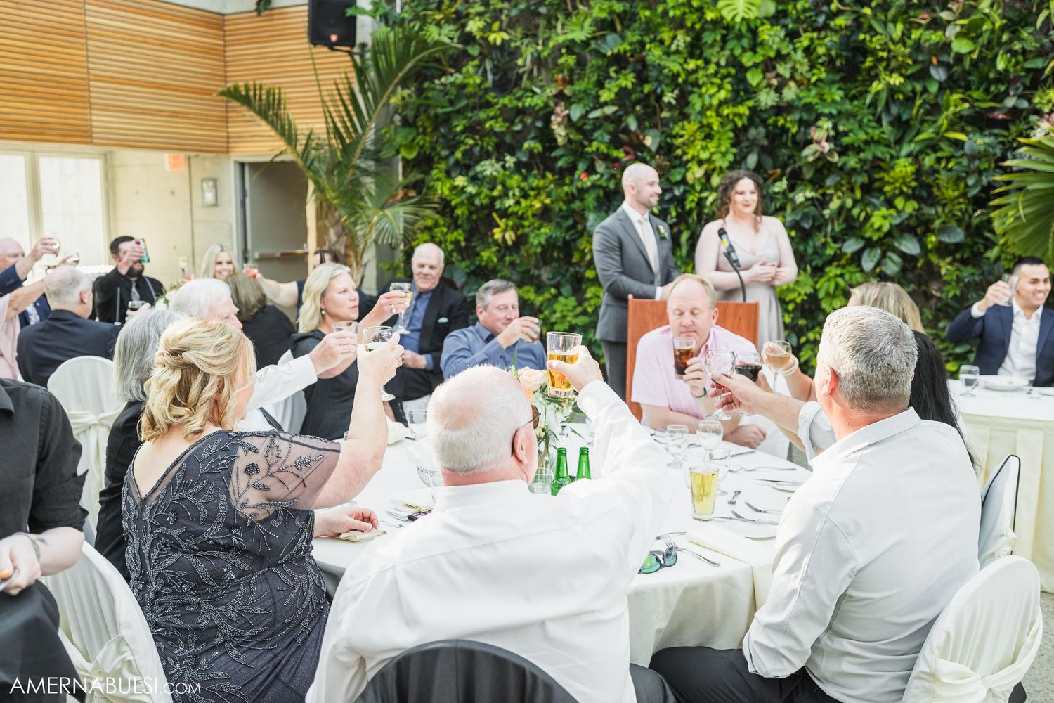 Guests seated at round tables during an elegant outdoor wedding reception with green wall backdrop.