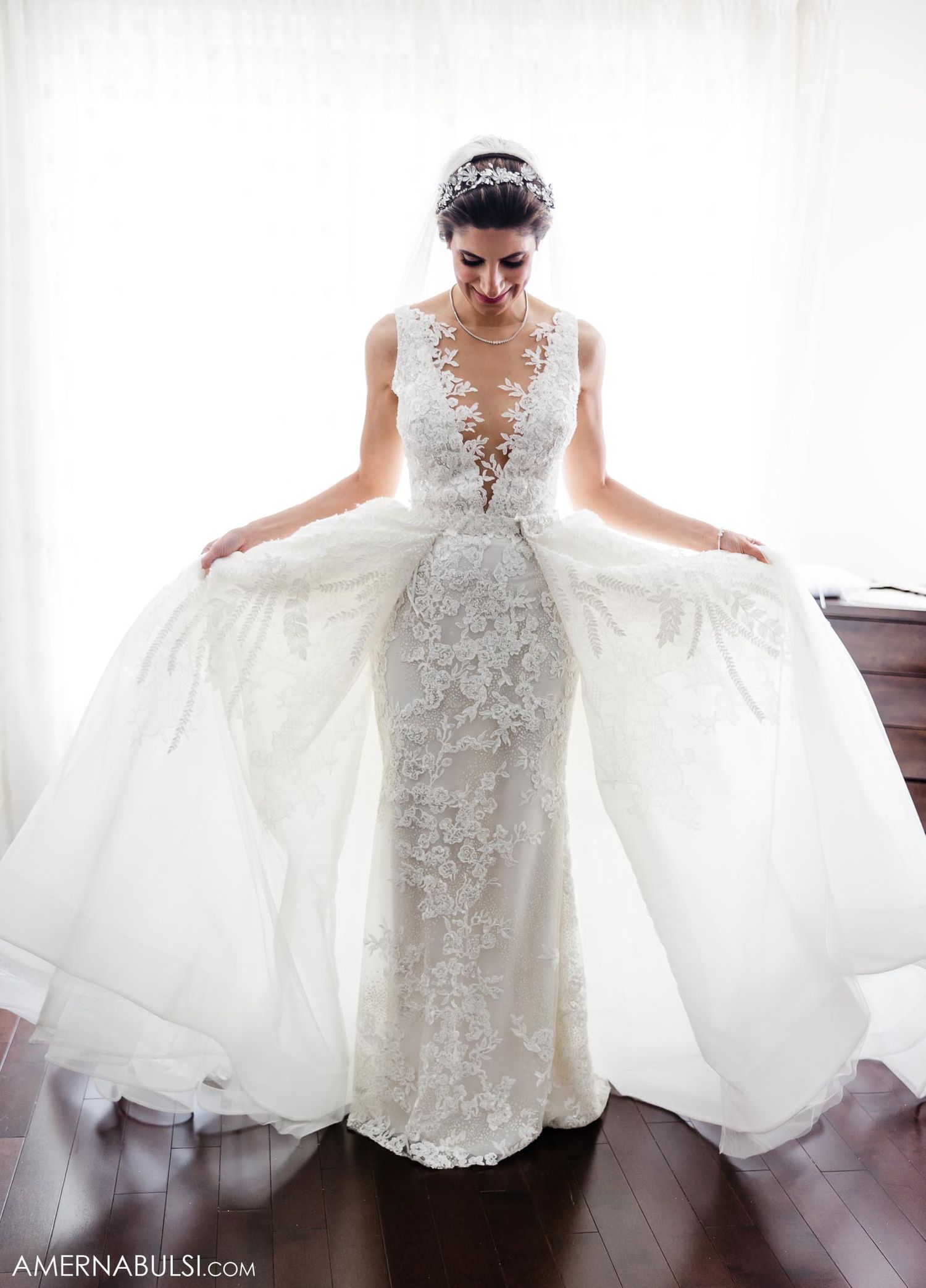 Bride showcases detailed lace wedding gown with flowing overlay in natural light.