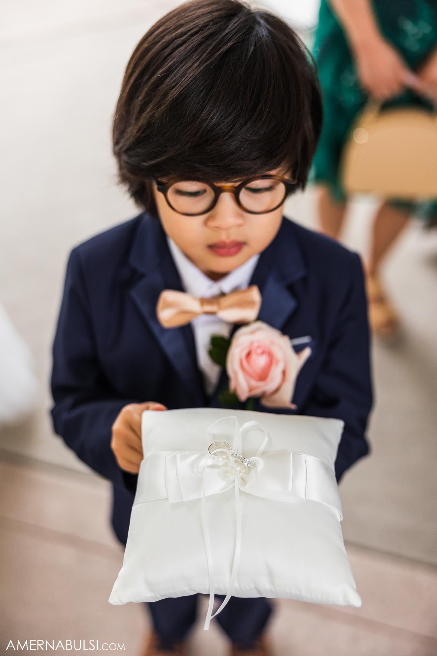 A ring bearer in a navy suit and bowtie holds a white satin pillow.