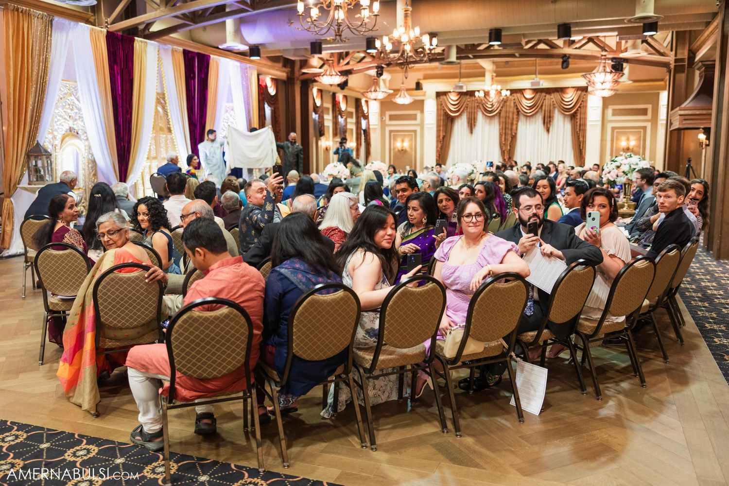 Guests seated in ornate banquet chairs fill an elegantly decorated wedding venue with warm lighting and drapes.