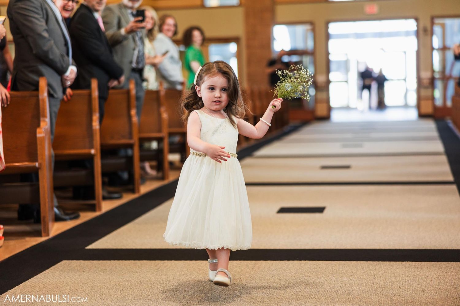 A young flower girl in a white dress walks down the church aisle during a wedding ceremony.