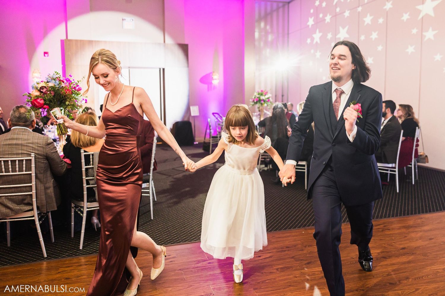 Wedding guests dance together with a flower girl in a modern reception hall with pink uplighting and twinkling lights.