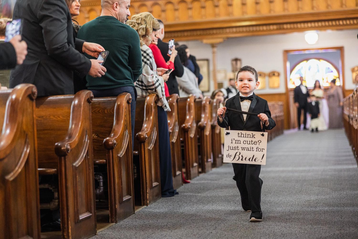 Young ring bearer in tuxedo walks down church aisle carrying wooden sign.