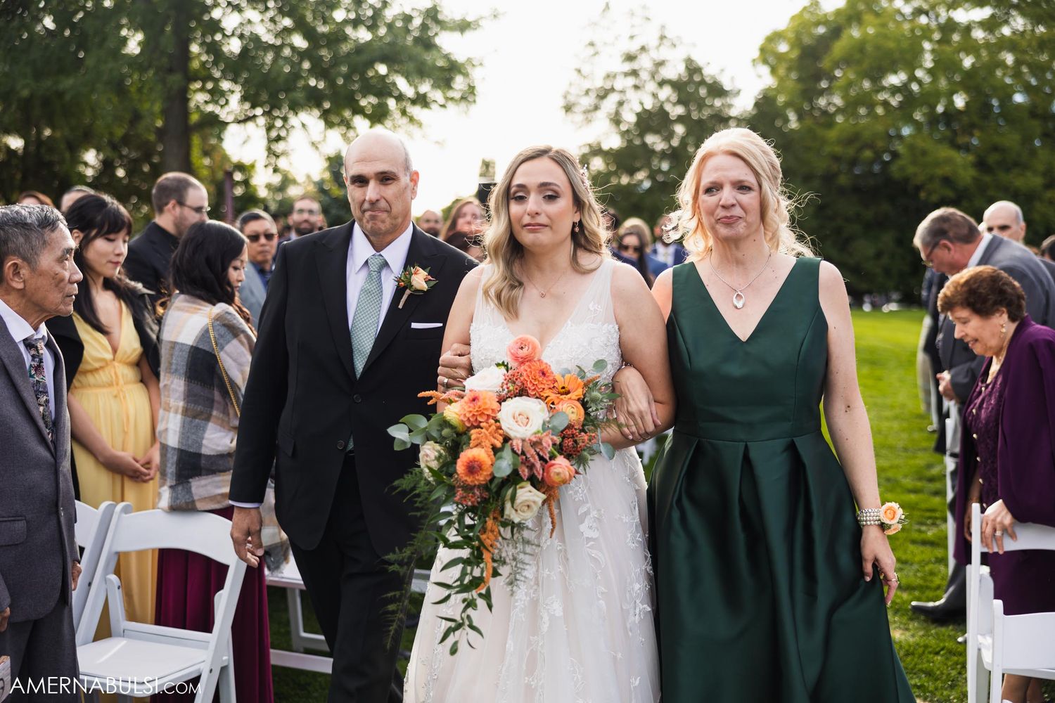 A wedding party walks down a grassy aisle with the bride in white and bridesmaid in emerald green.