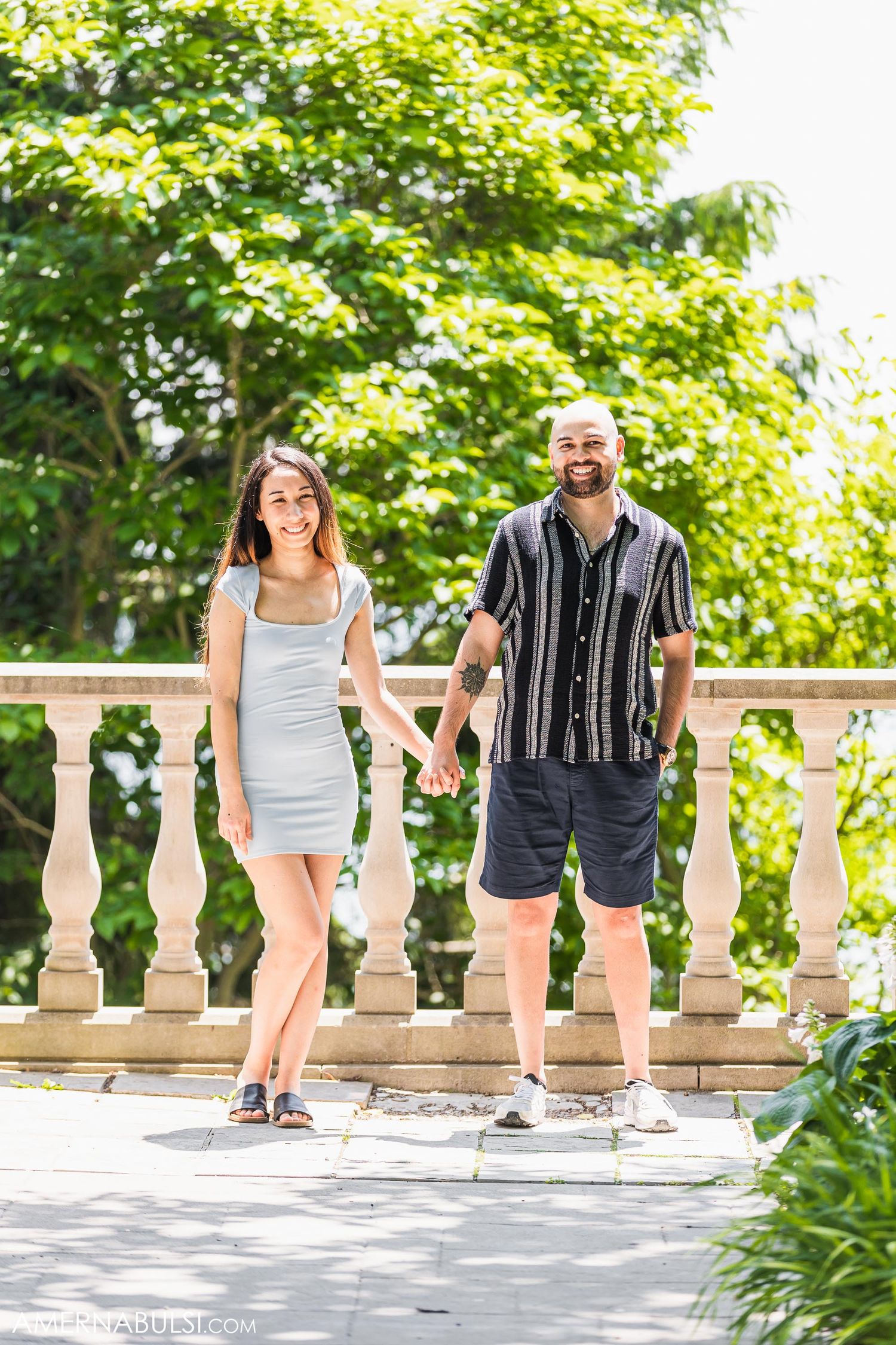 Happy couple holding hands while standing on a stone balcony surrounded by lush green trees.