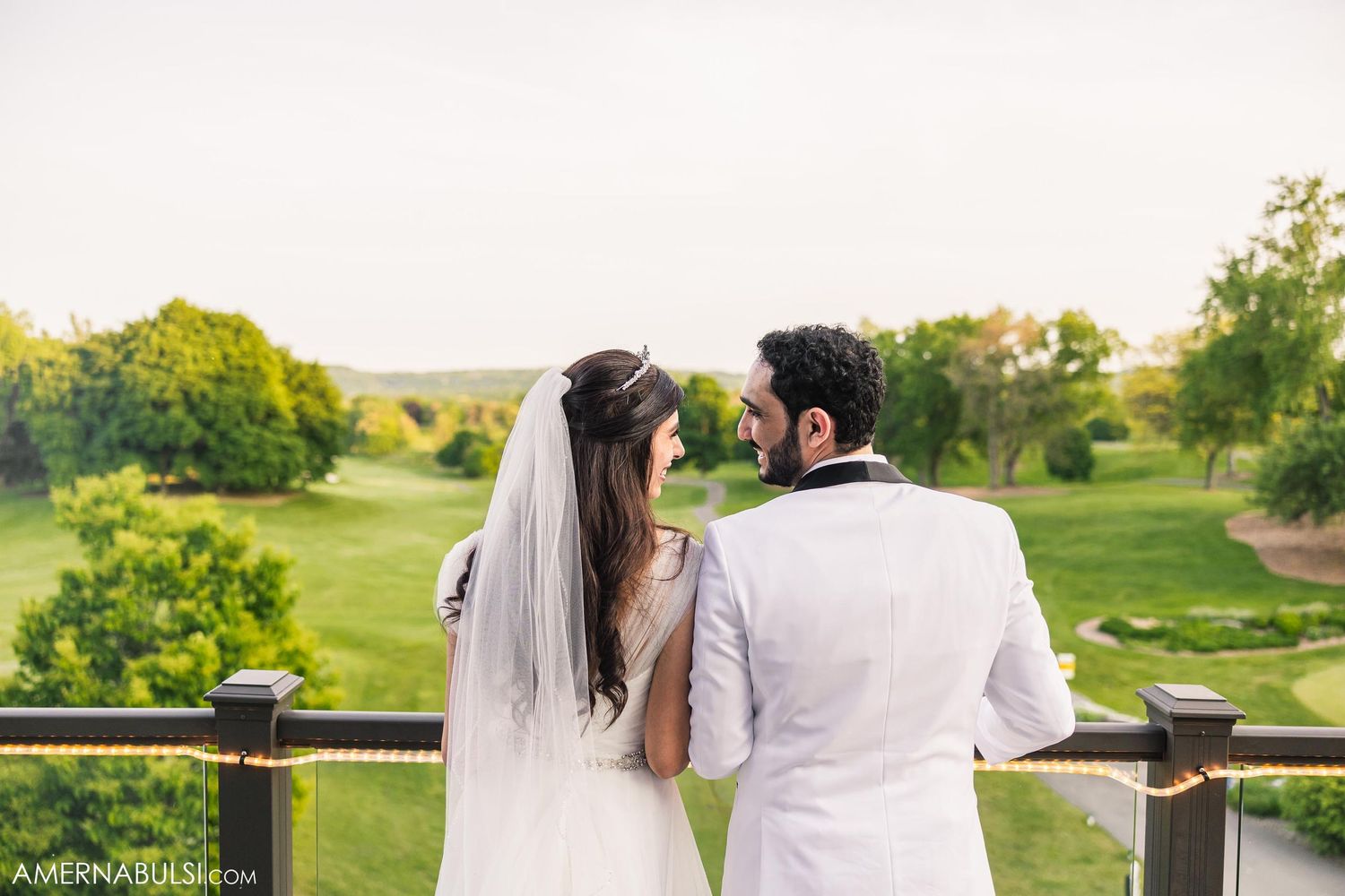 A couple in wedding attire looking at each other on a balcony overlooking green landscape.