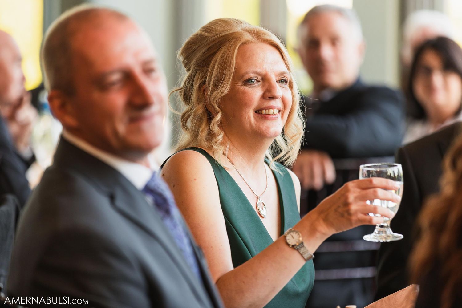 Wedding guest in emerald dress smiles while holding champagne glass at reception.