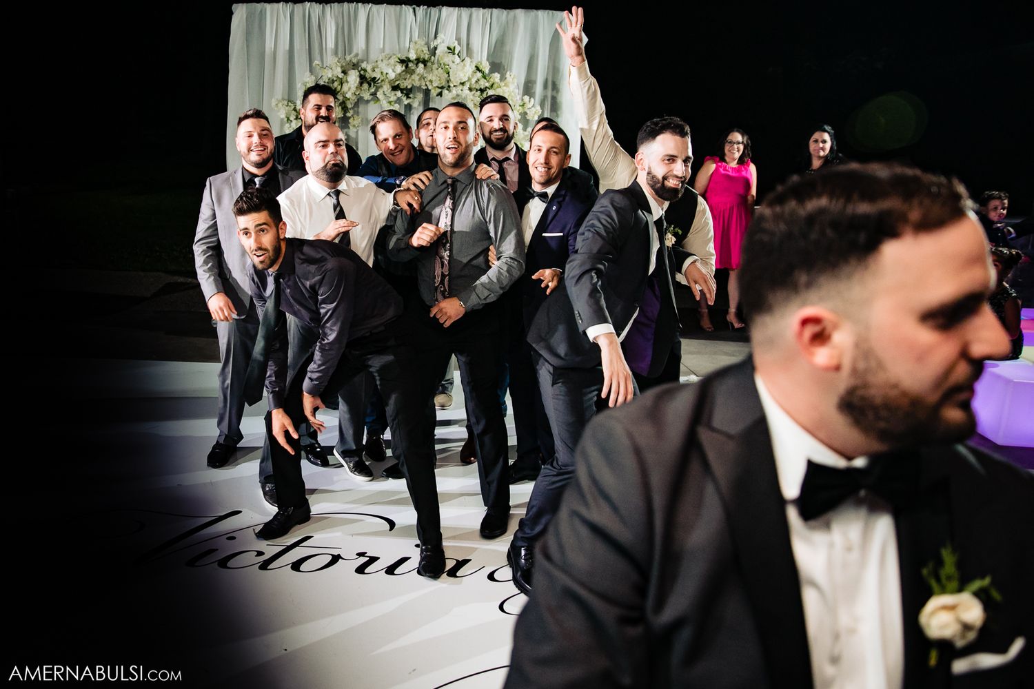 Wedding guests dance energetically on a dark dance floor during an evening reception celebration.
