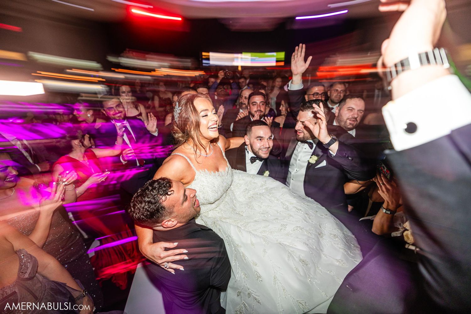 Wedding reception crowd dancing and celebrating under vibrant red and purple lights.