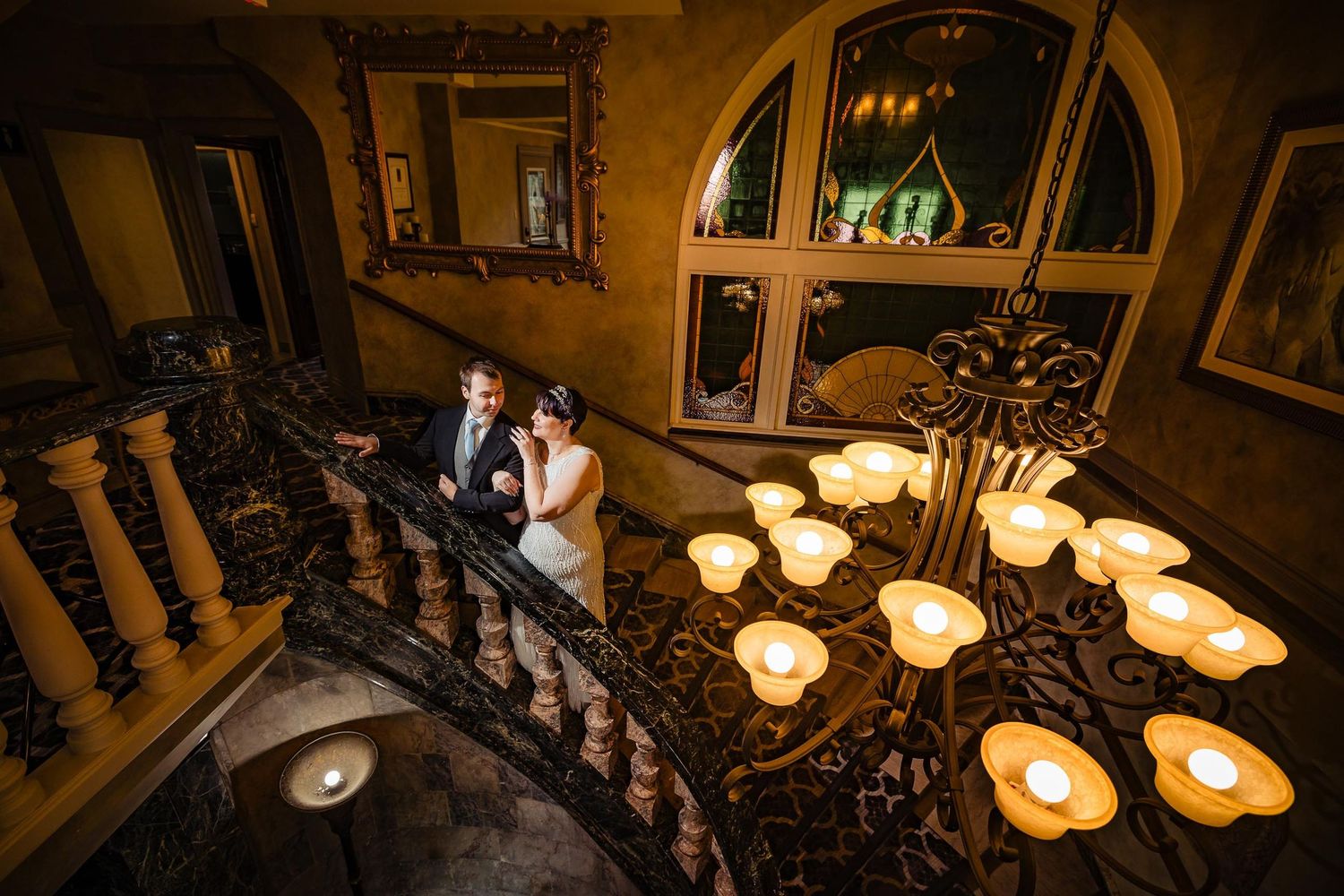 Romantic couple pose on ornate staircase illuminated by vintage-style wall sconces.