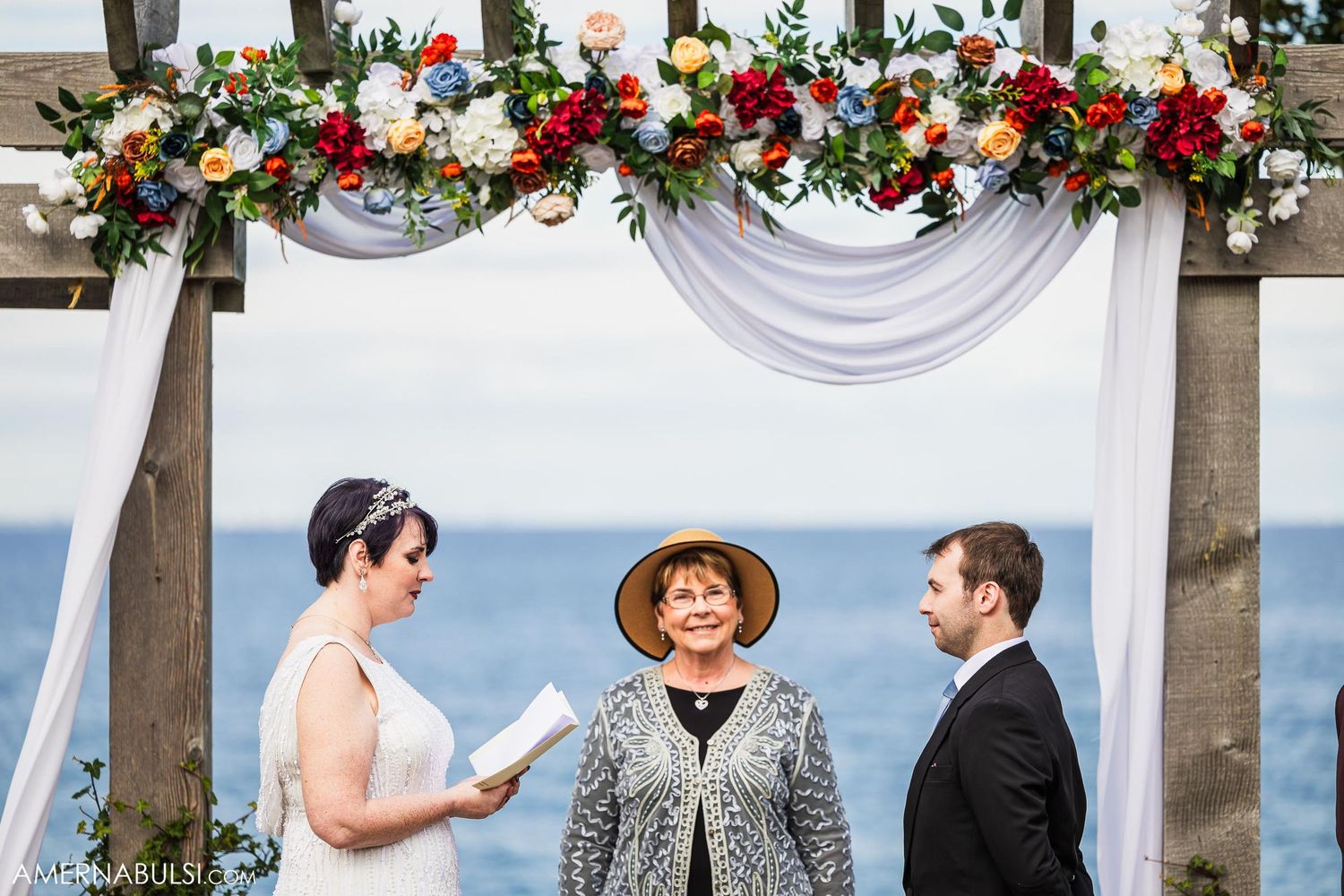 Wedding ceremony under wooden pergola decorated with colorful flowers and white fabric.