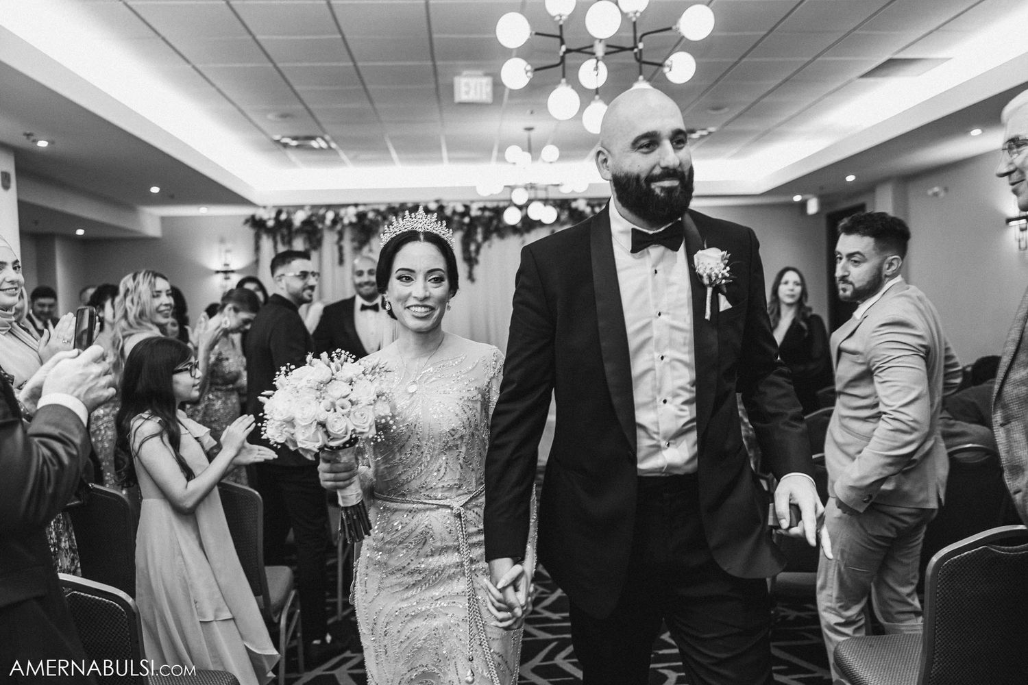 Wedding couple walking down aisle while guests celebrate in reception hall with chandelier.
