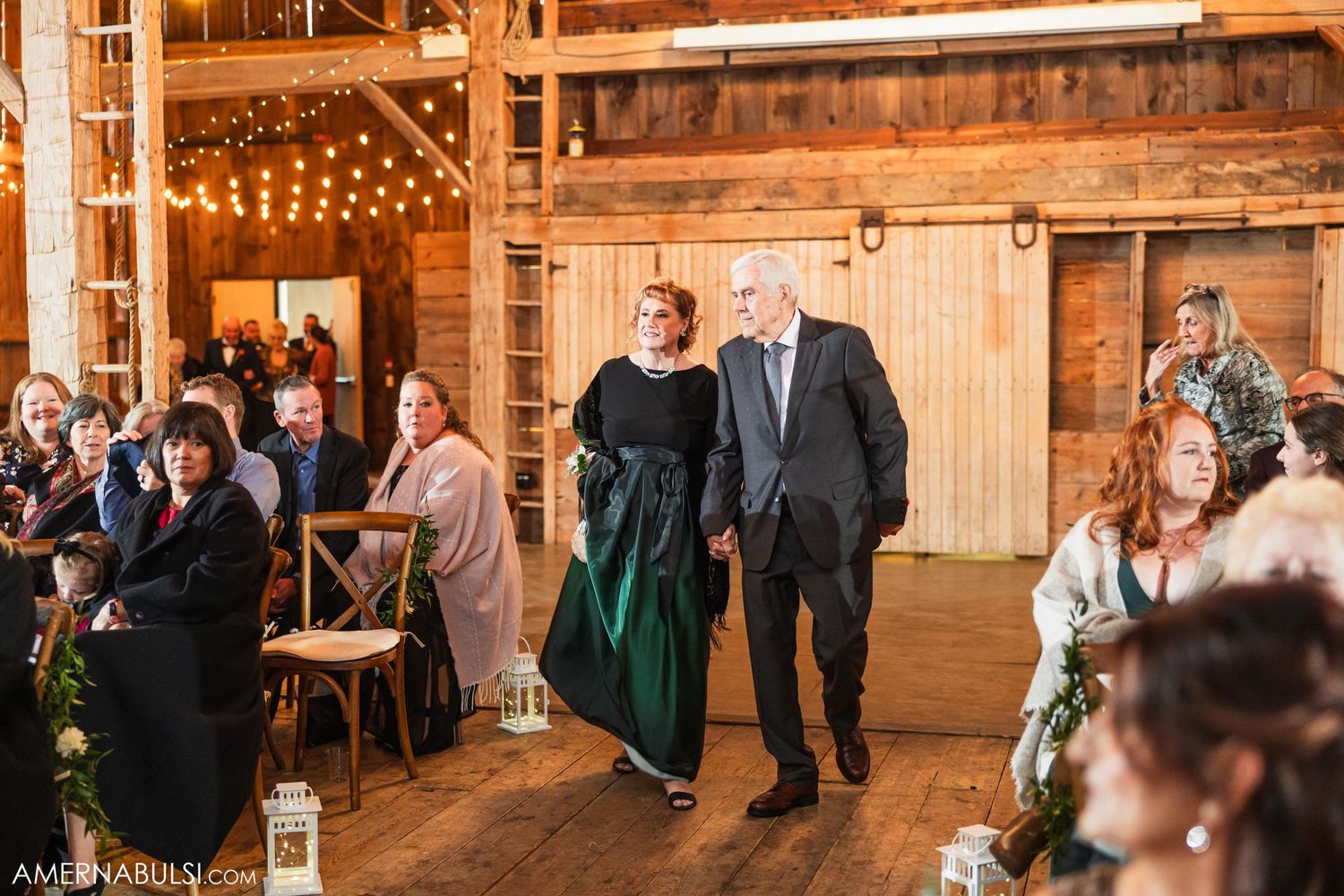 Wedding party members walk down aisle in rustic barn venue decorated with string lights.