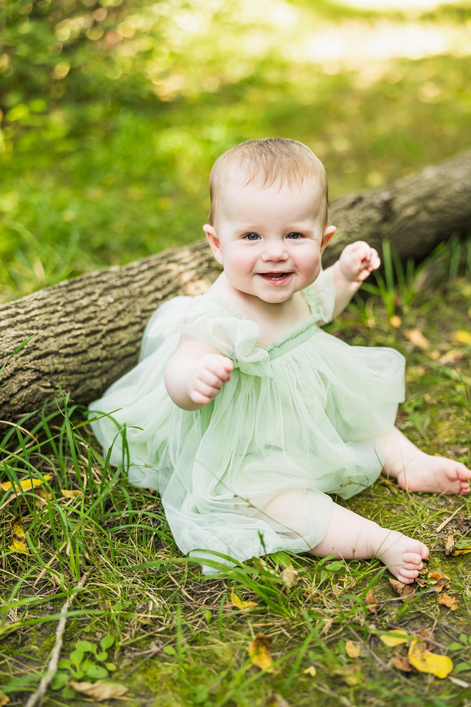 Happy baby in a mint green dress sits on grass near a fallen log in dappled sunlight.