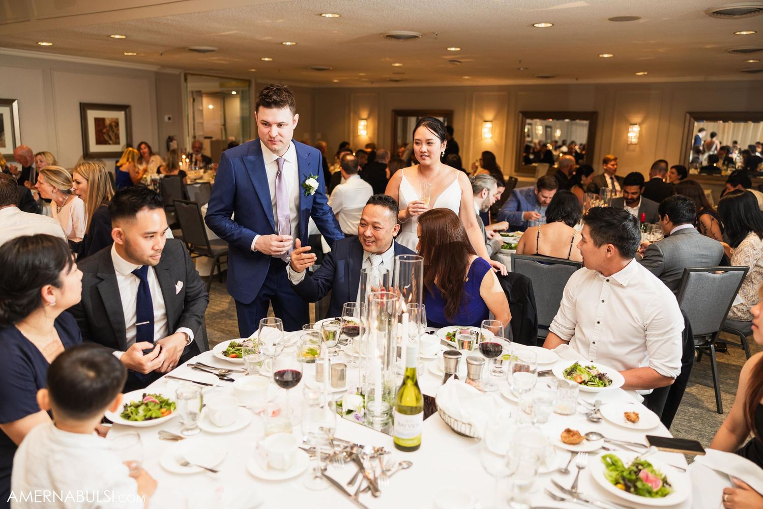 Wedding reception guests gather around elegantly set dinner table with wine and formal place settings.