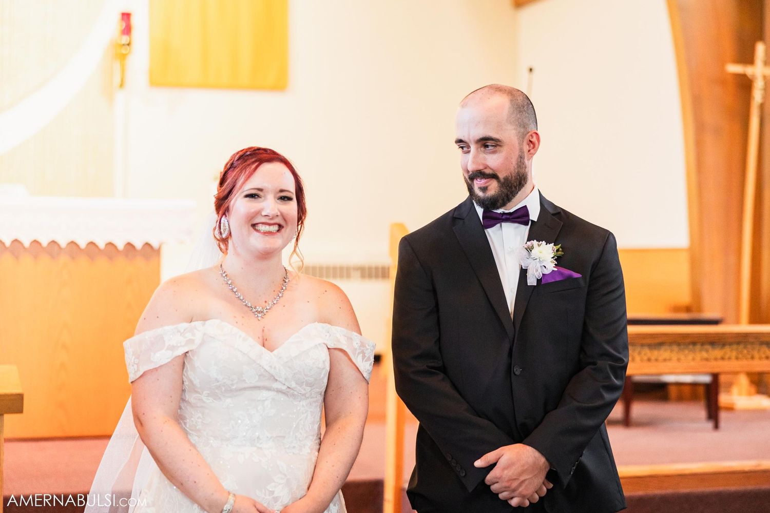 Bride in off-shoulder gown and groom in tuxedo standing at church altar.