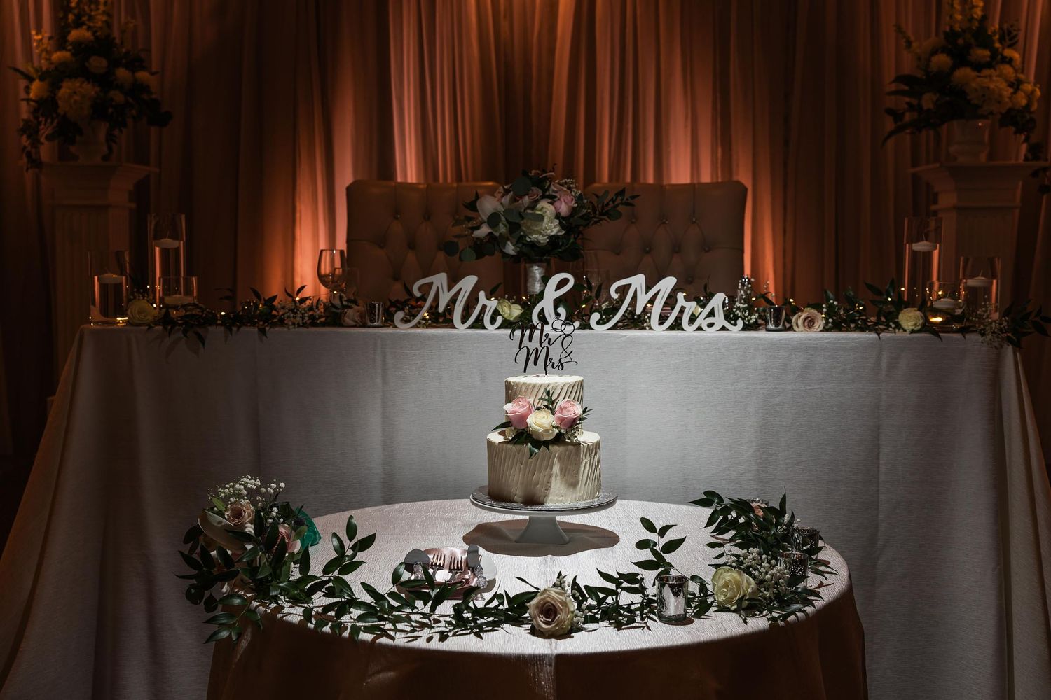 Wedding cake table with Mr & Mrs sign and romantic uplighting in background.