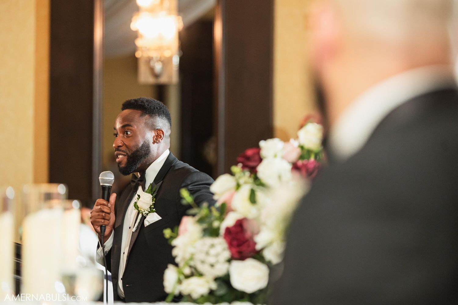 A joyful speaker gives a toast at an elegant wedding reception with floral centerpieces and ambient lighting.