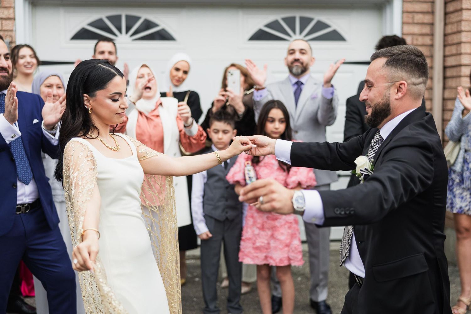 A couple performs a traditional dance while guests cheer in front of a brick house.