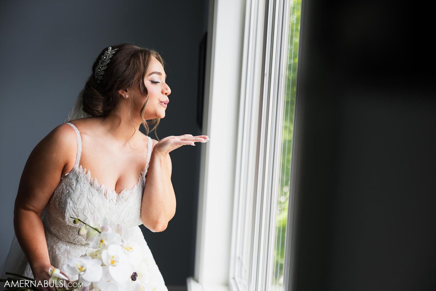 Person in white wedding dress blowing kiss by window in romantic bridal portrait.