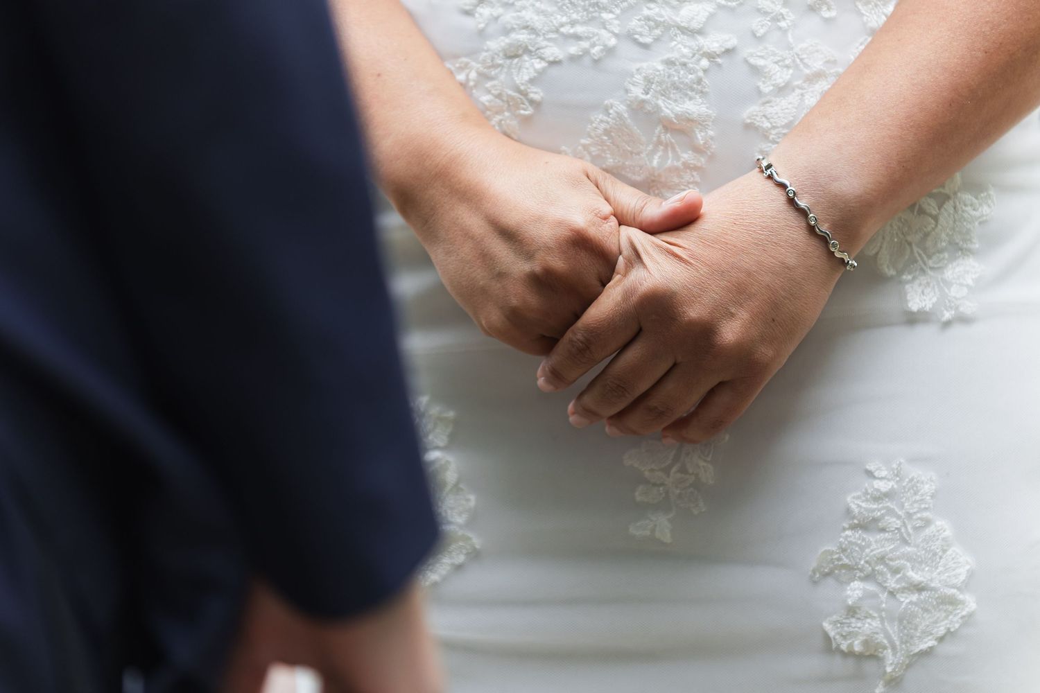 Close-up of bride and groom holding hands, bride wearing lace wedding dress and delicate bracelet.