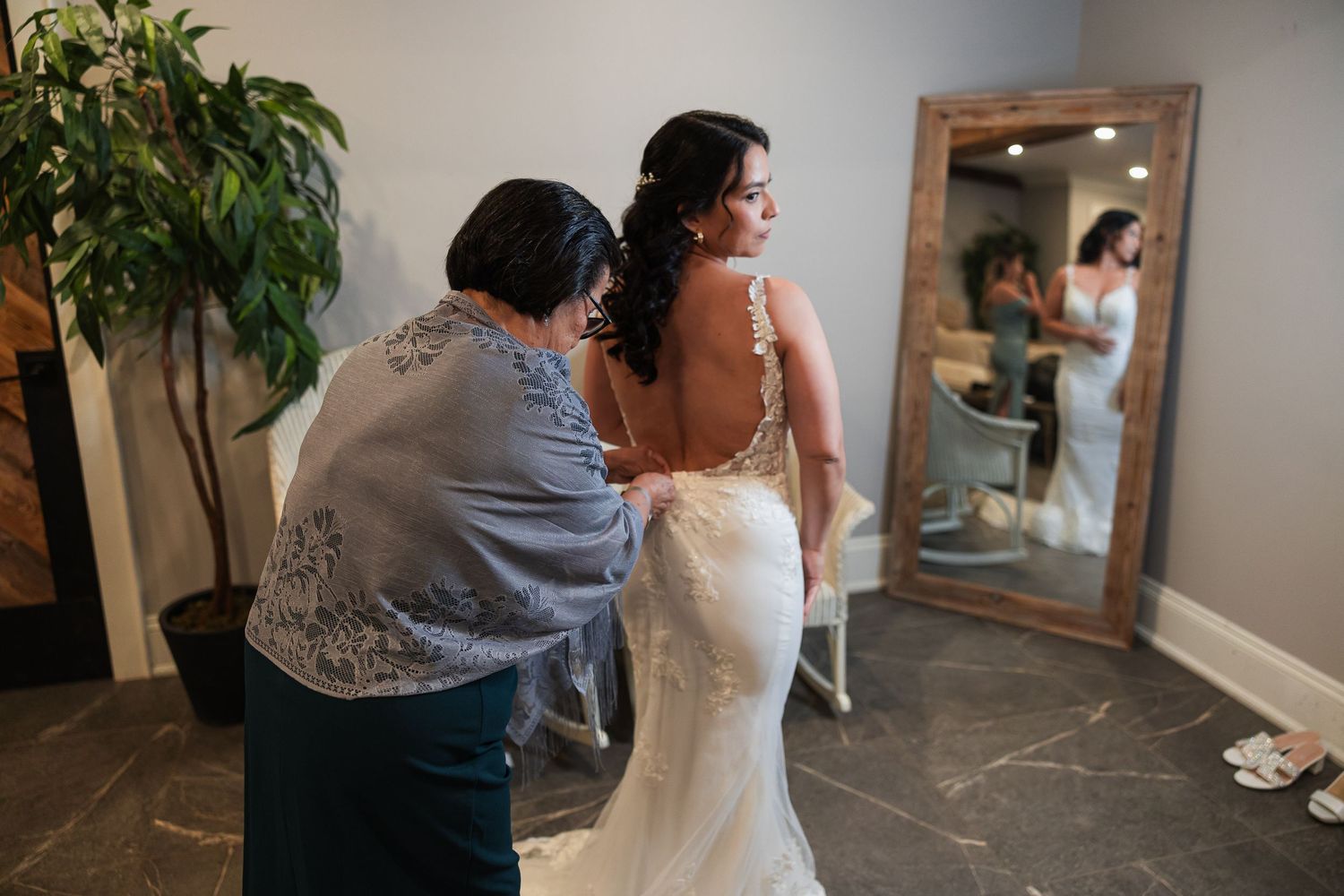 A bride getting help with her wedding dress from a family member in a bridal suite, with a mirror reflection visible.