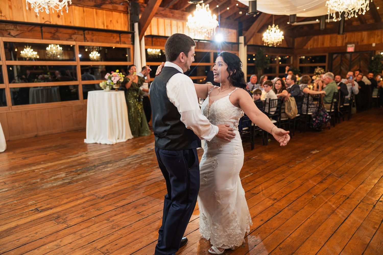 Bride and groom share their first dance on a wooden dance floor in a rustic barn wedding venue with chandeliers.