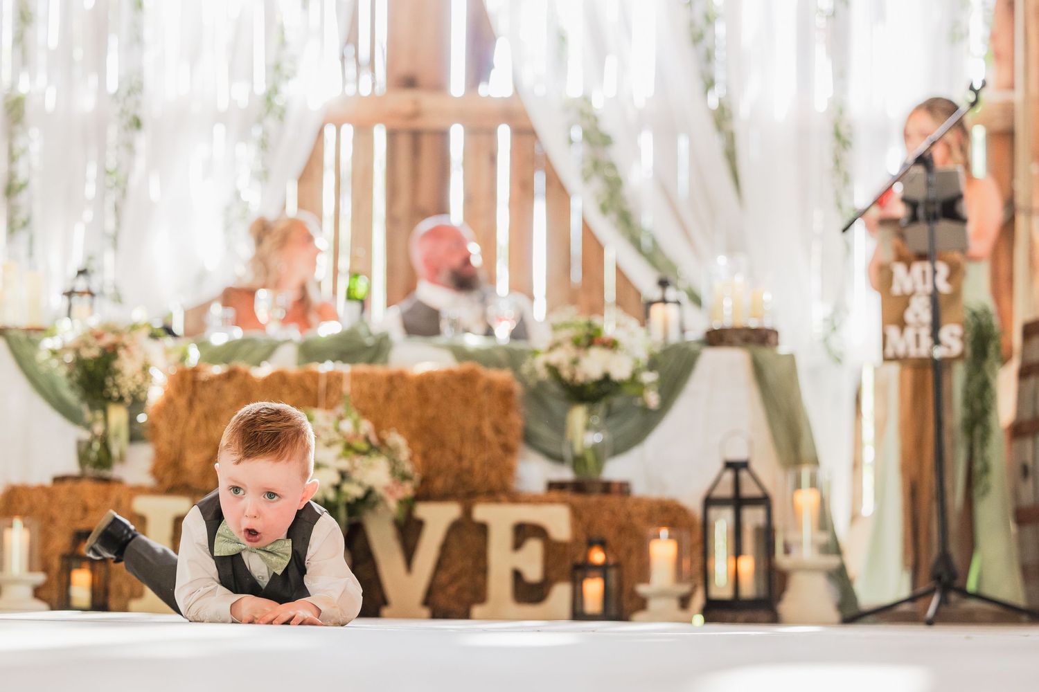 Young boy in vest rests on table at rustic barn wedding reception with Mr & Mrs sign and hay bales.