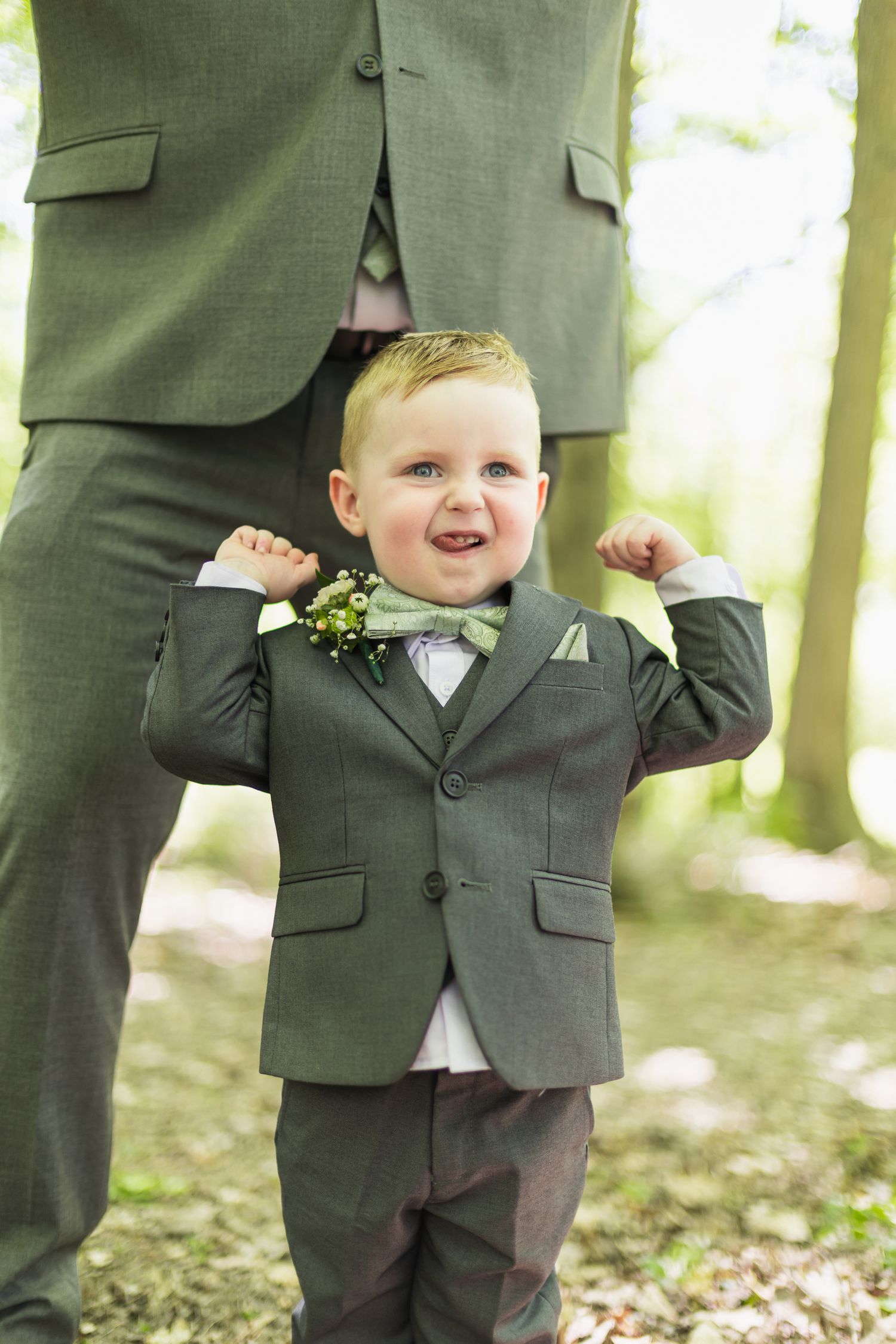 Adorable toddler boy in matching green suit and bow tie stands proudly between adult legs at a wedding.