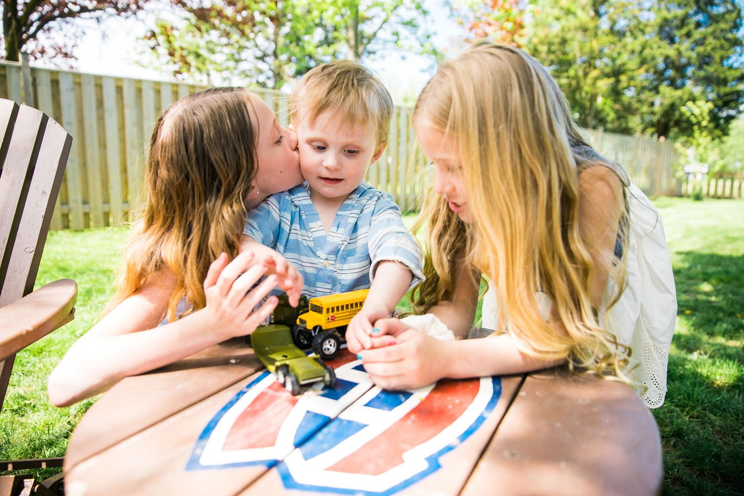 A group of children gather around a table outdoors to paint with watercolors on a sunny day.