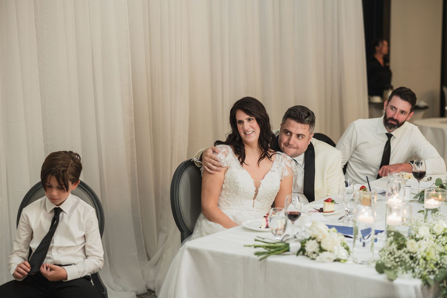 Bride and groom seated at wedding reception head table with guests, white floral centerpieces and candles.