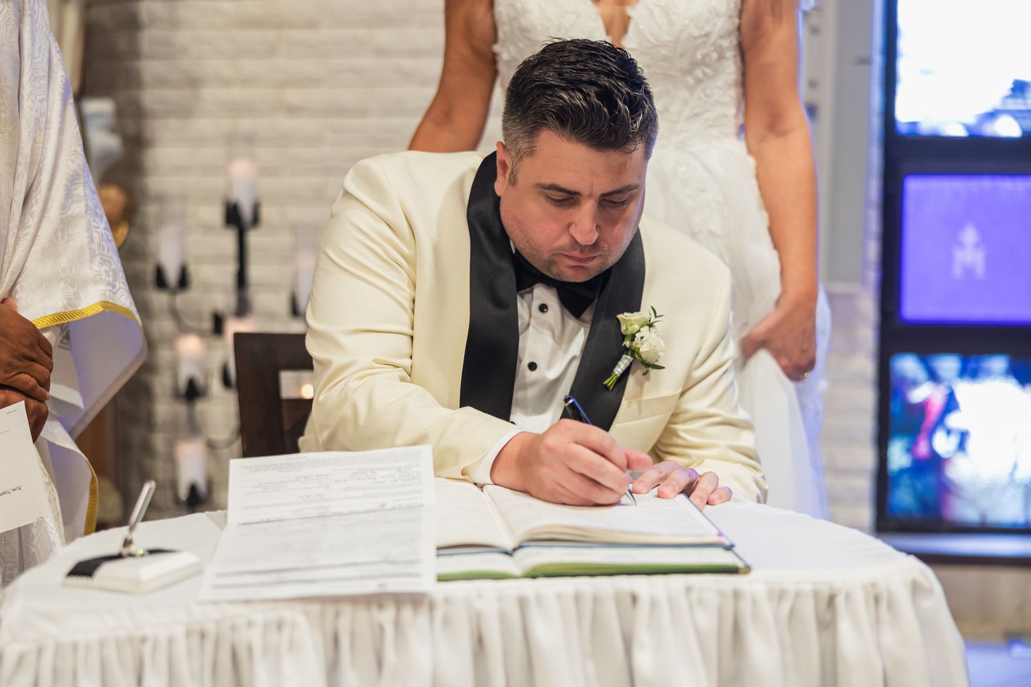 Groom in white tuxedo signing wedding registry at decorated table during ceremony.