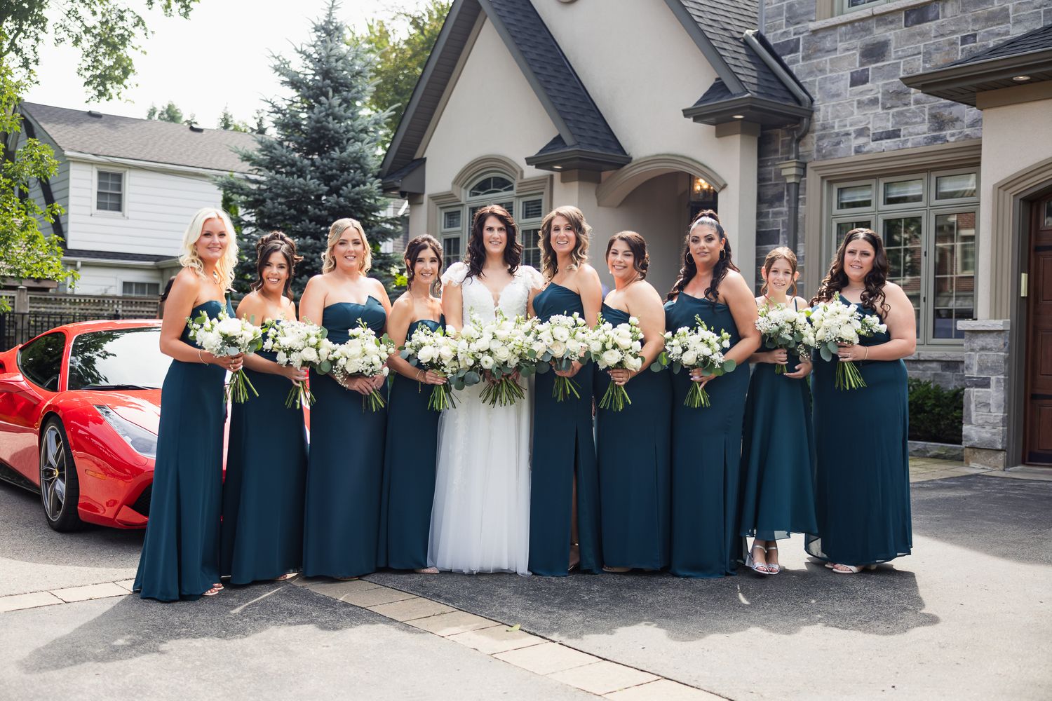Bride with bridesmaids in teal gowns holding white bouquets pose outdoors near a red Ferrari.