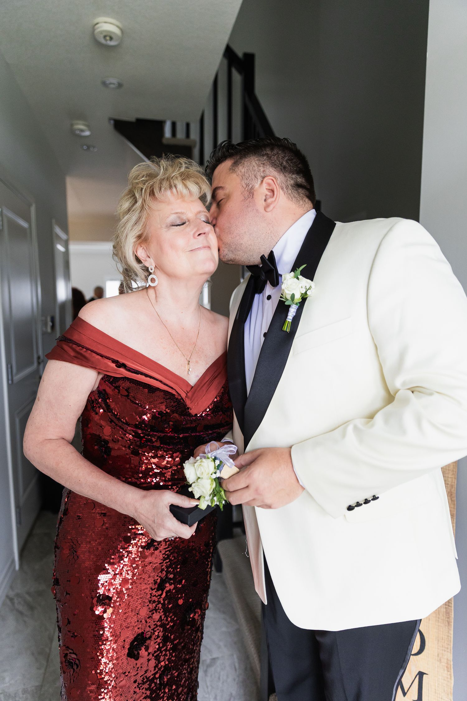 Groom in white tuxedo kissing mother in red sequin dress on cheek before wedding ceremony.