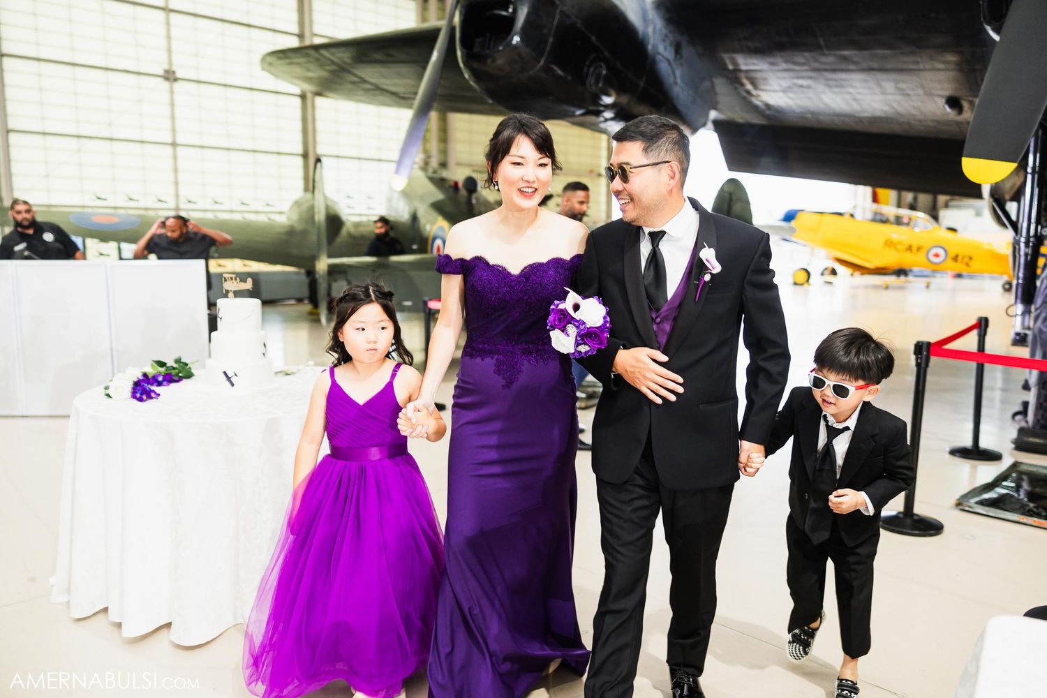 Wedding party in purple and black attire poses in aircraft hangar.