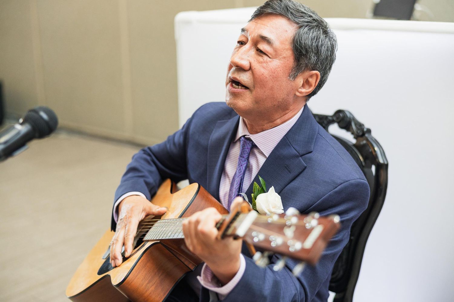 A performer in a navy suit plays acoustic guitar during a wedding celebration while seated.