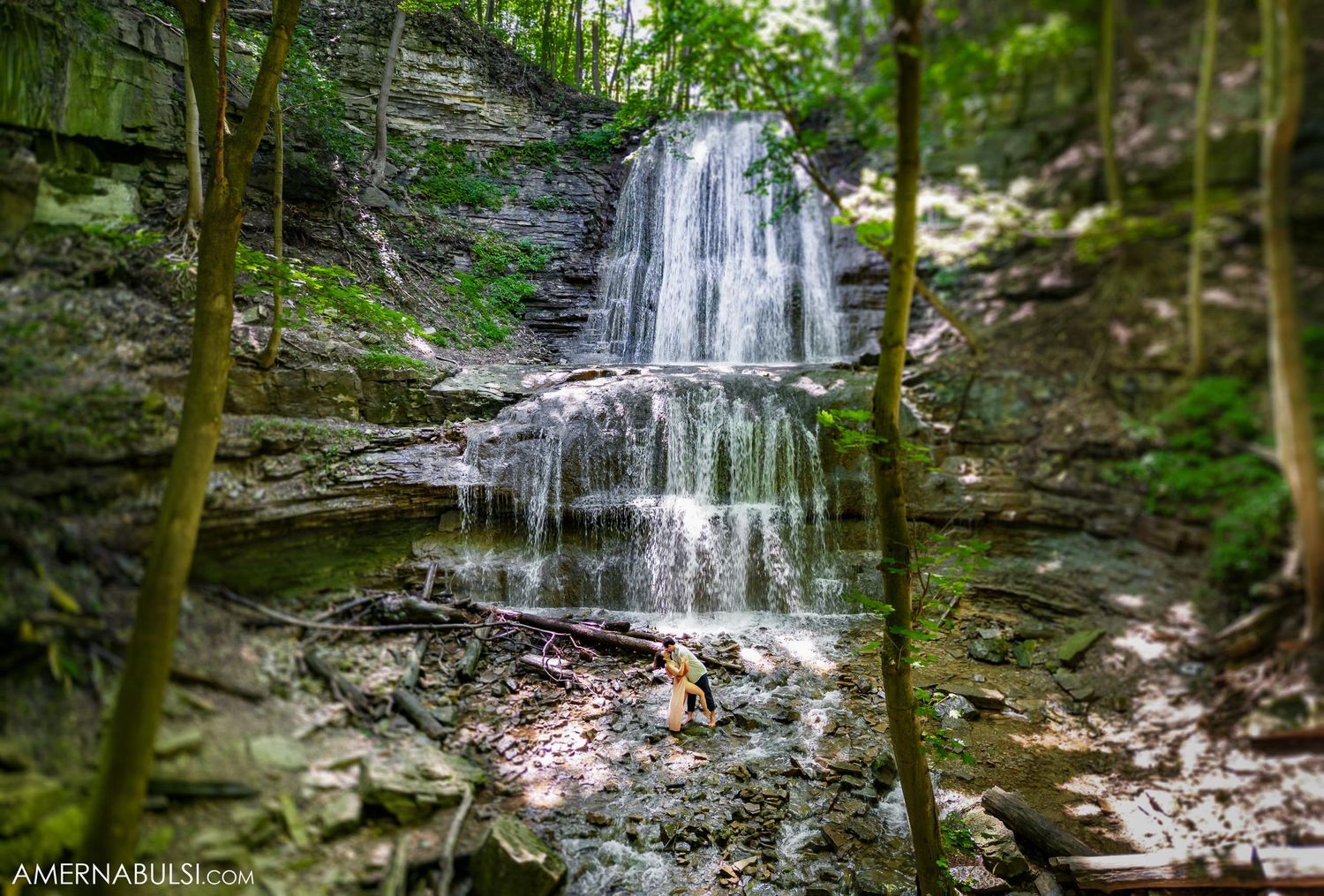 A serene multi-tiered waterfall cascades down mossy rocks in a lush green forest setting.