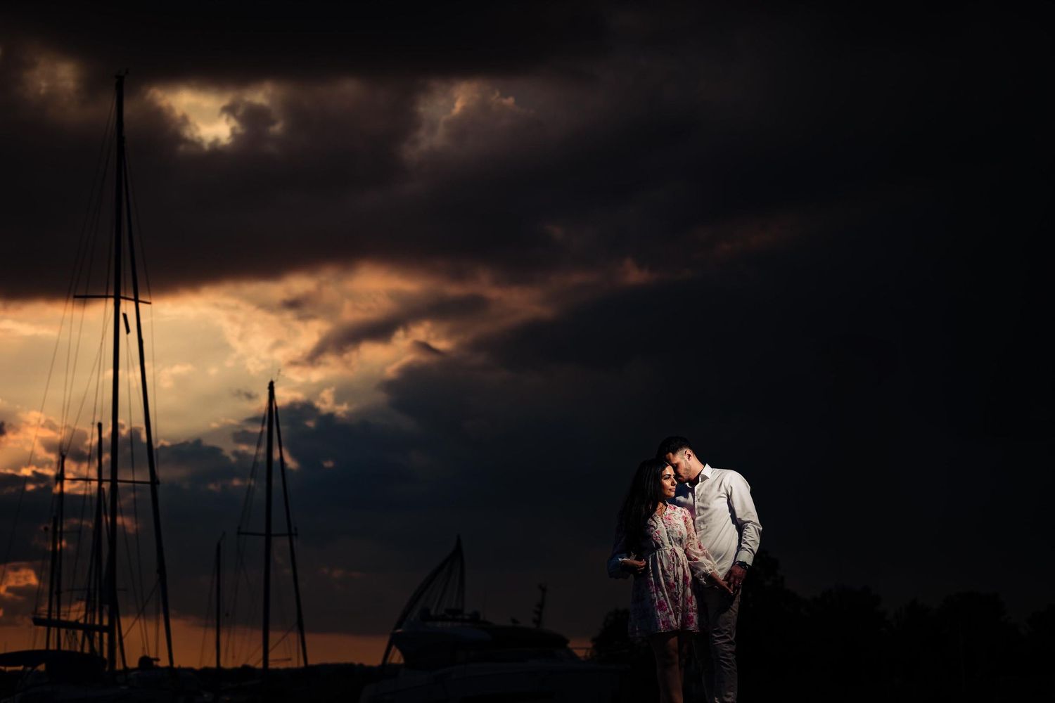 Dramatic sunset silhouettes couple against marina with storm clouds.