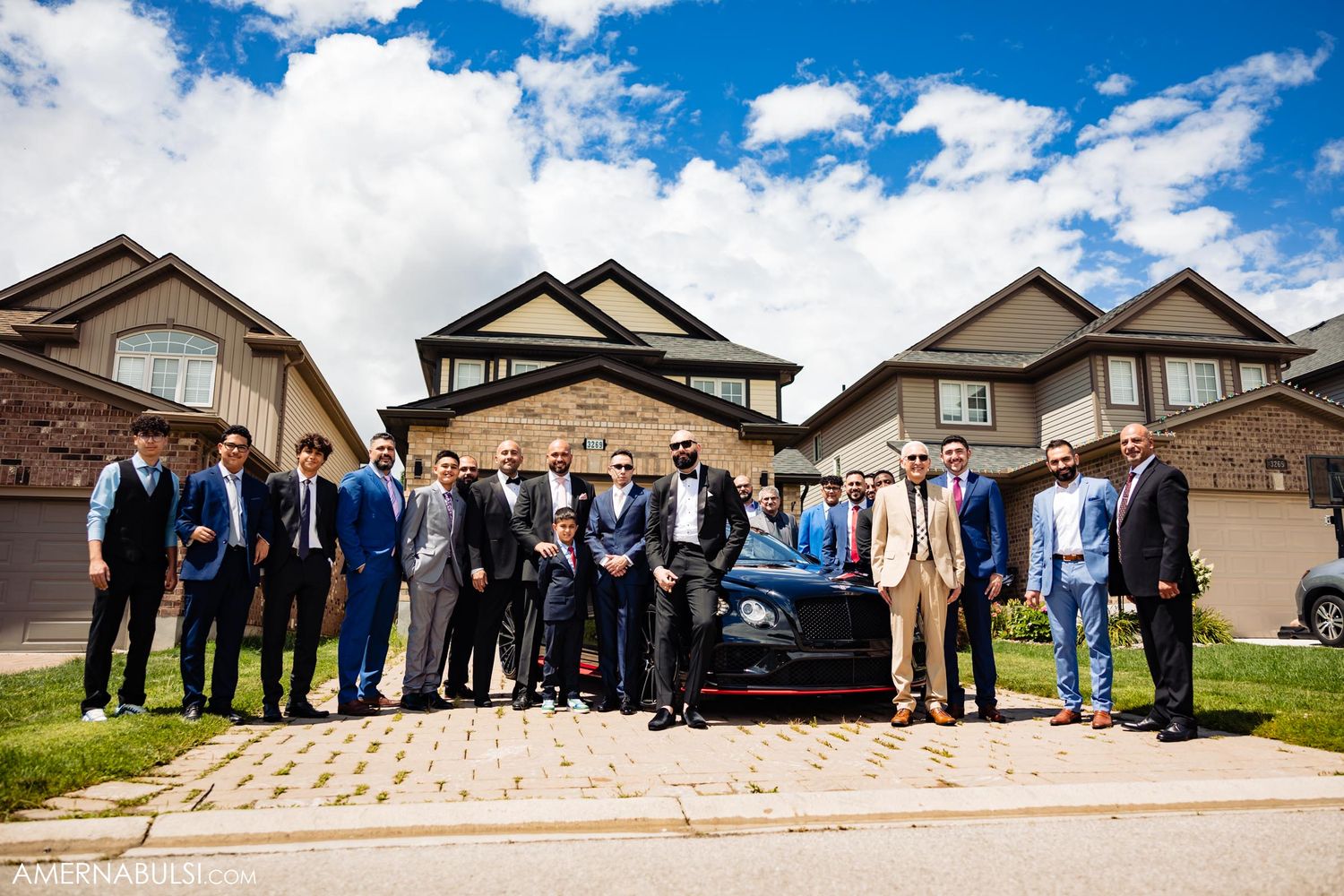 Wedding party poses with luxury car in front of suburban homes under blue sky.