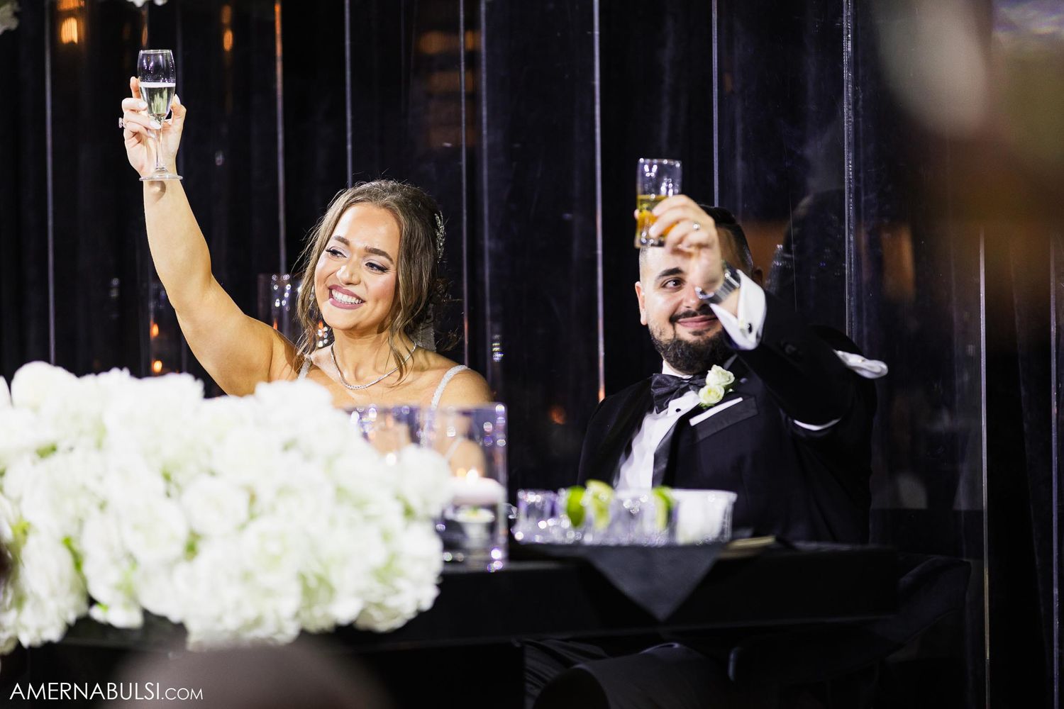 A couple raises champagne glasses during an elegant wedding toast with white flowers.