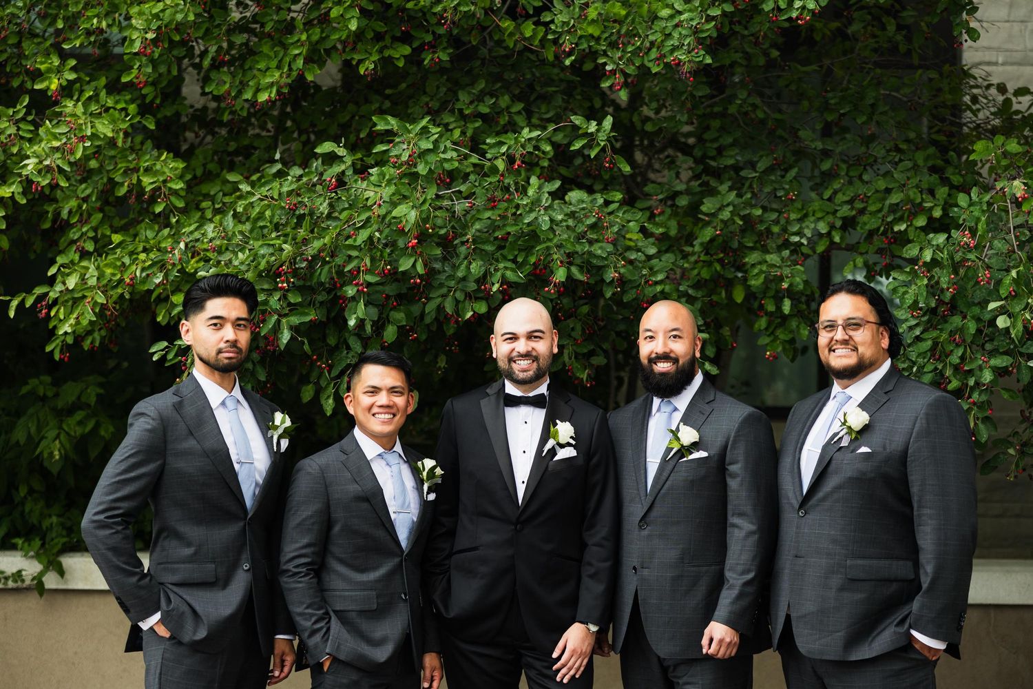 Groomsmen in dark suits pose together outdoors against green foliage background.