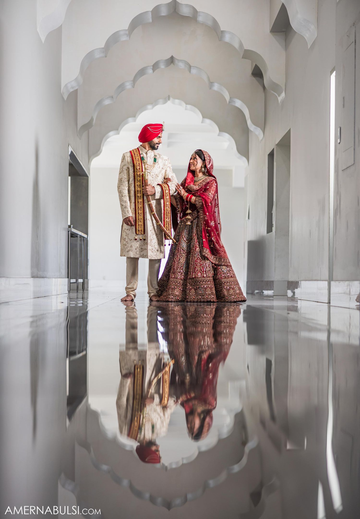 Couple in traditional Indian wedding attire standing under ornate white archway with reflection on polished floor.
