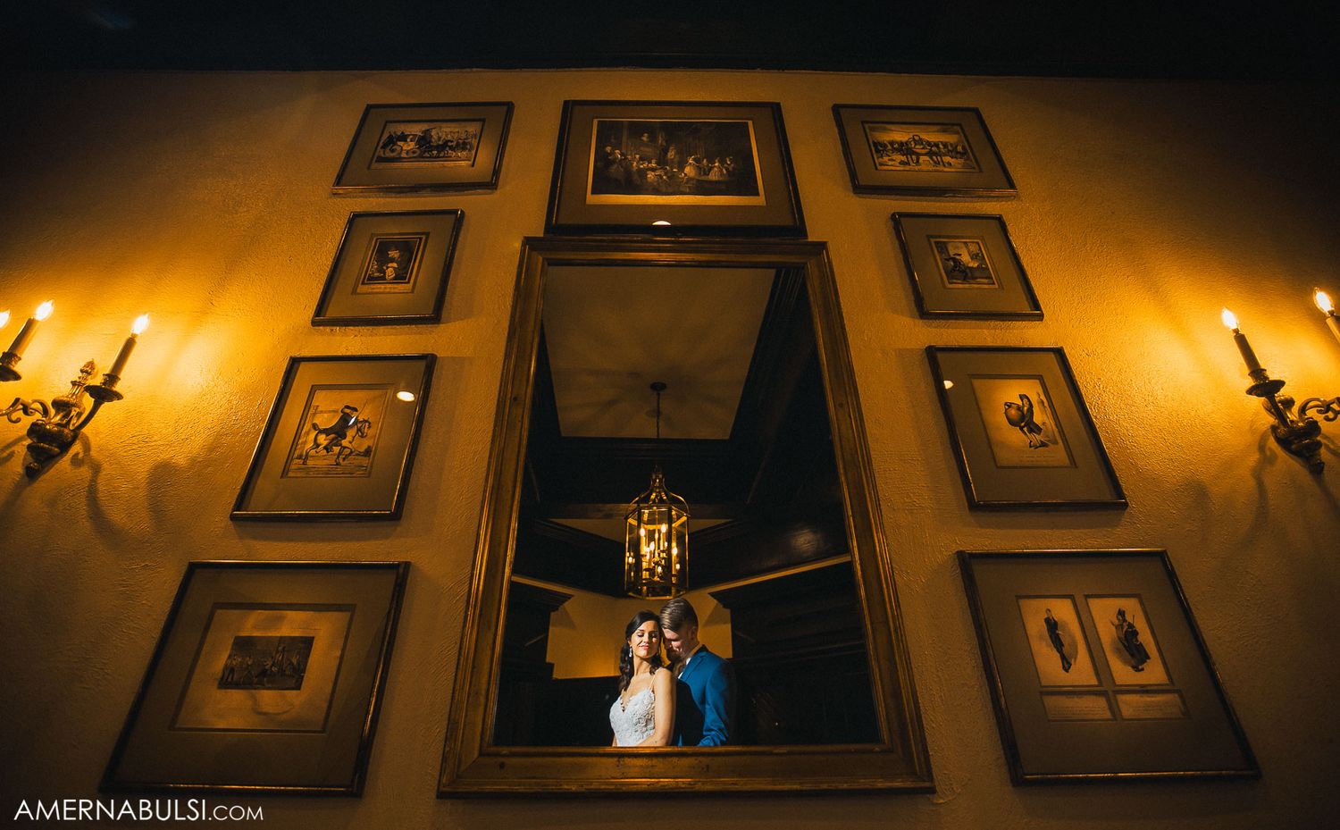 Dramatic wedding portrait reflected in ornate mirror surrounded by framed photos and wall sconces.